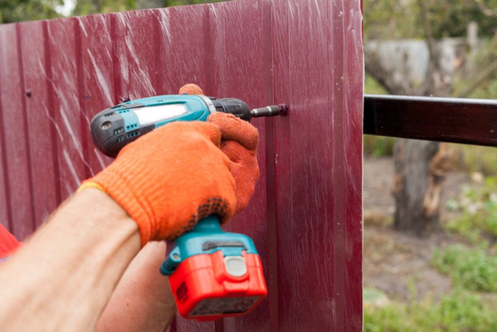 A person using an electric drill to paint the side of a fence.