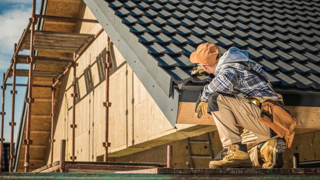 A man in work clothes and boots is on the roof of a house.